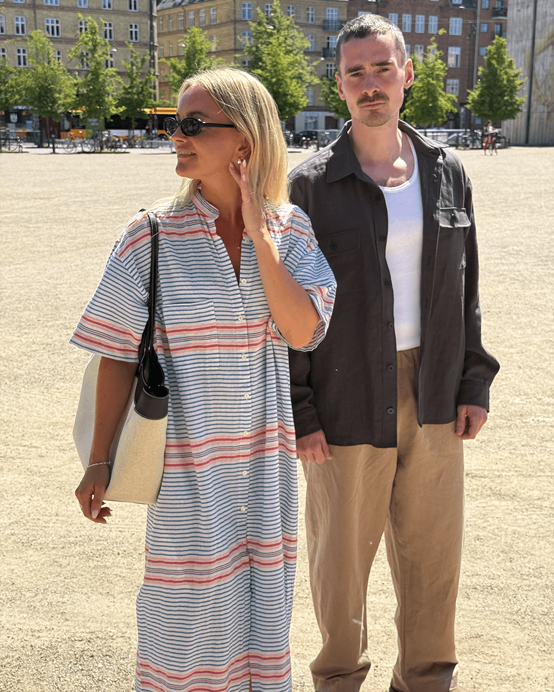 Two stylish individuals outdoors, one in a striped big shirt dress and the other in a dark button-up shirt.
