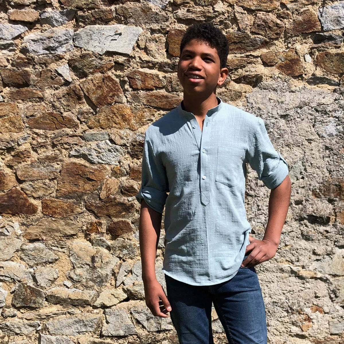 Young model showcasing the Men's Tunic paper pattern in a light blue fabric against a stone wall.
