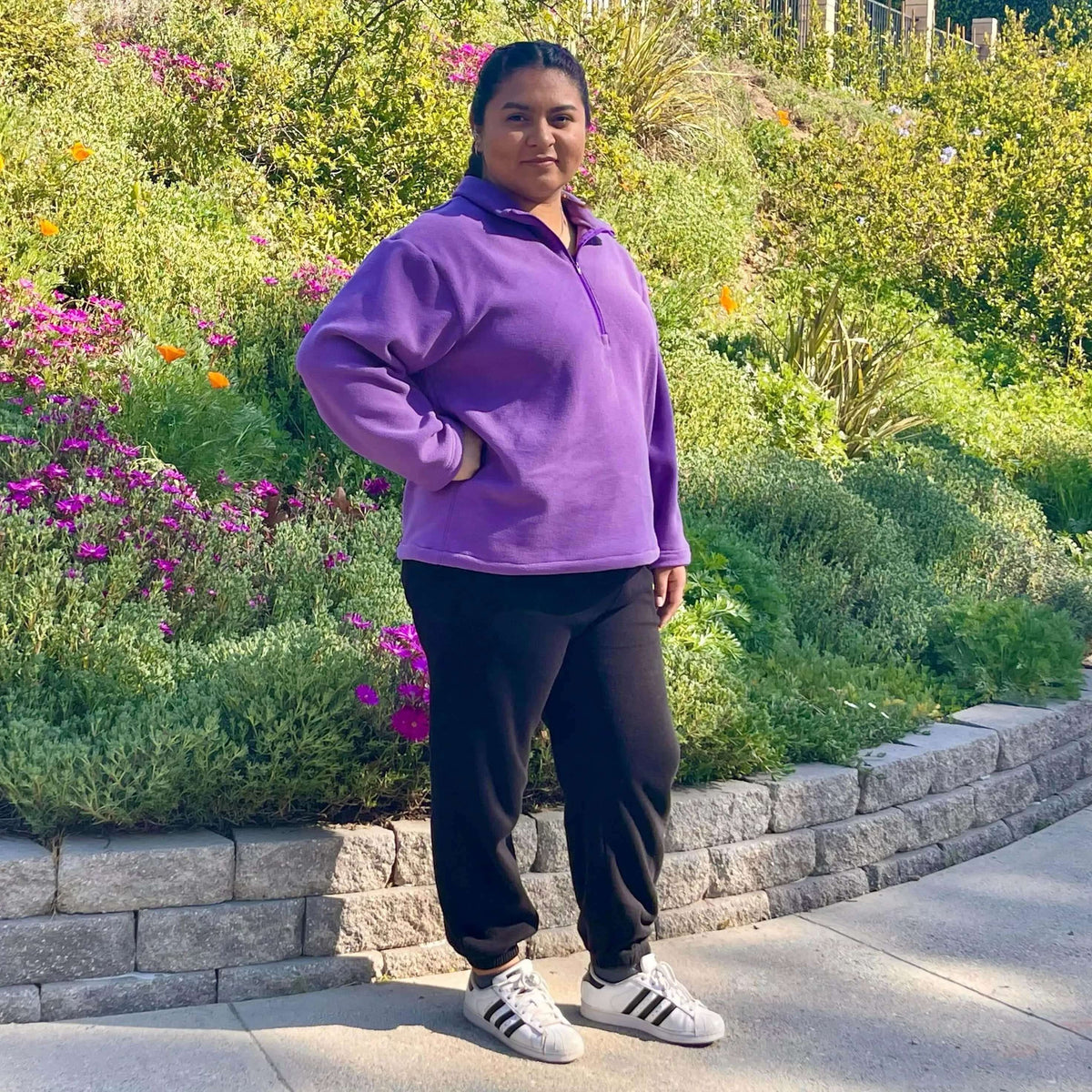 Model wearing a Half-Zipper Sweater Paper Pattern in purple, paired with black pants, surrounded by vibrant flowers.