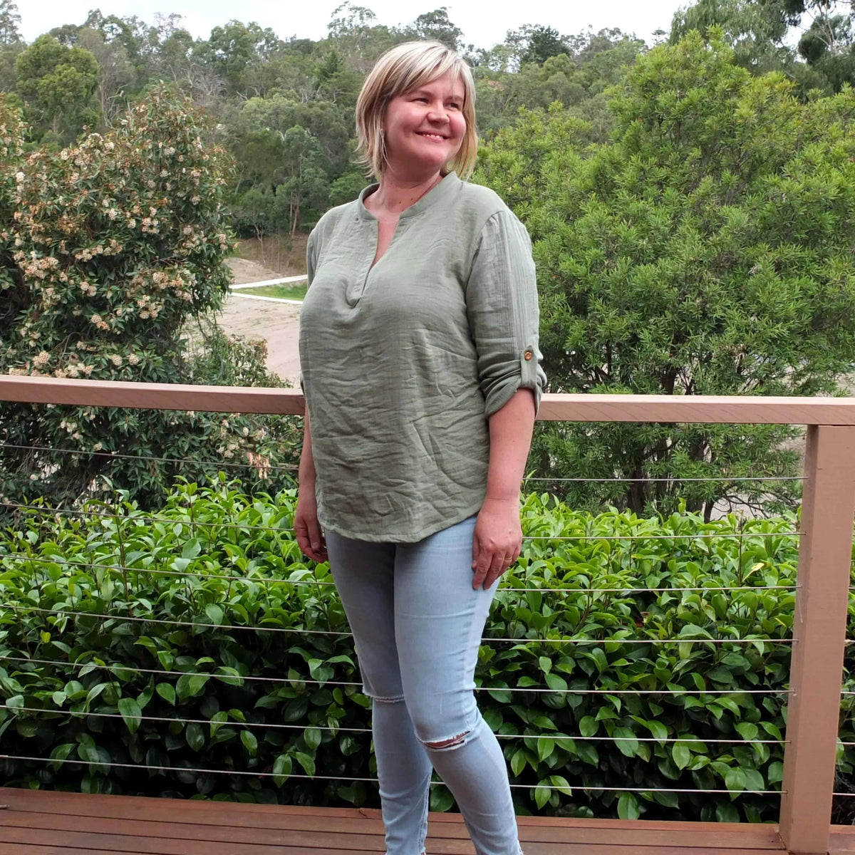 A woman wearing a relaxed Perfect Tunic Paper Pattern top with rolled sleeves, standing outdoors in a green setting.