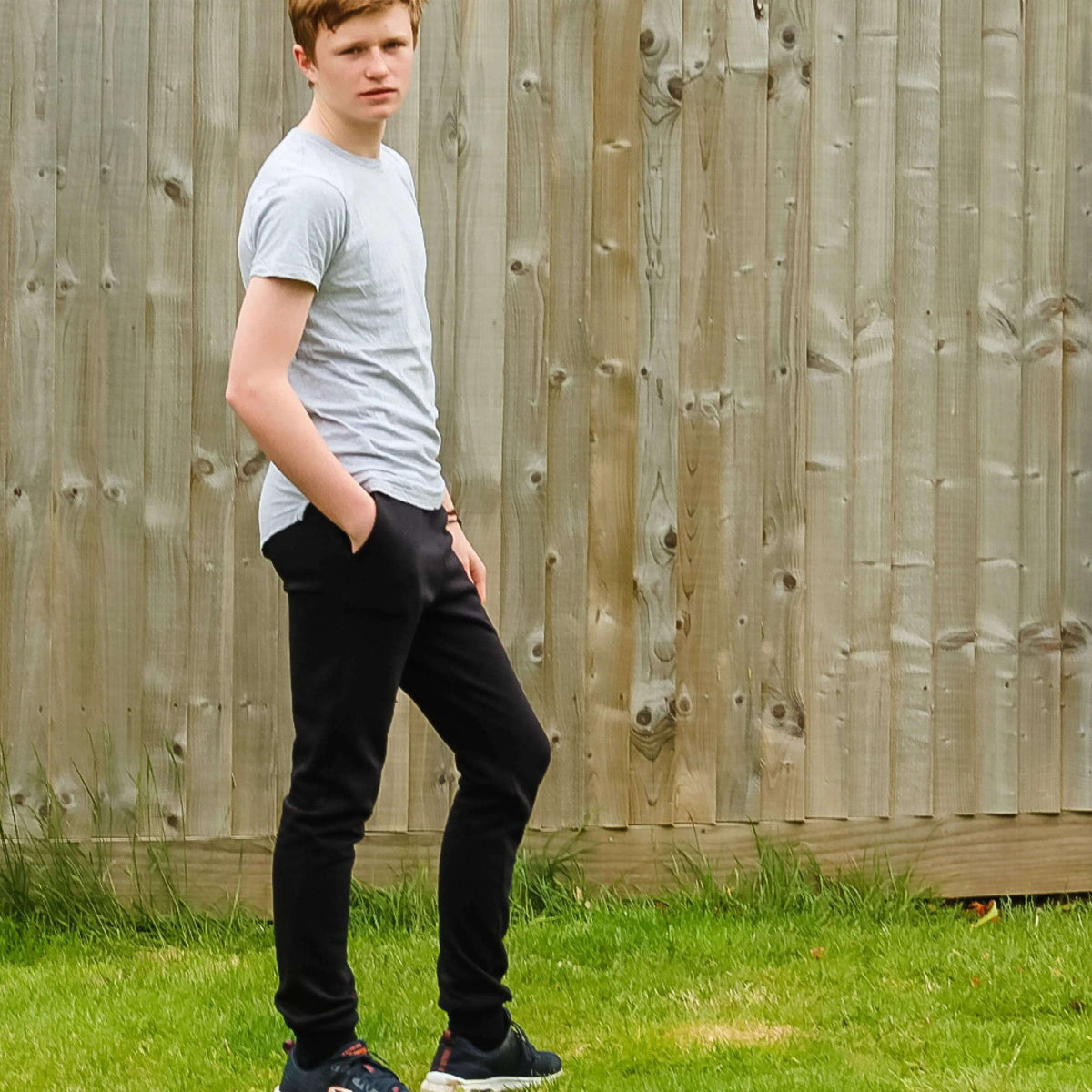 A young man modeling the comfortable Rebel Sweatpants Paper pattern against a wooden fence in a grassy area.