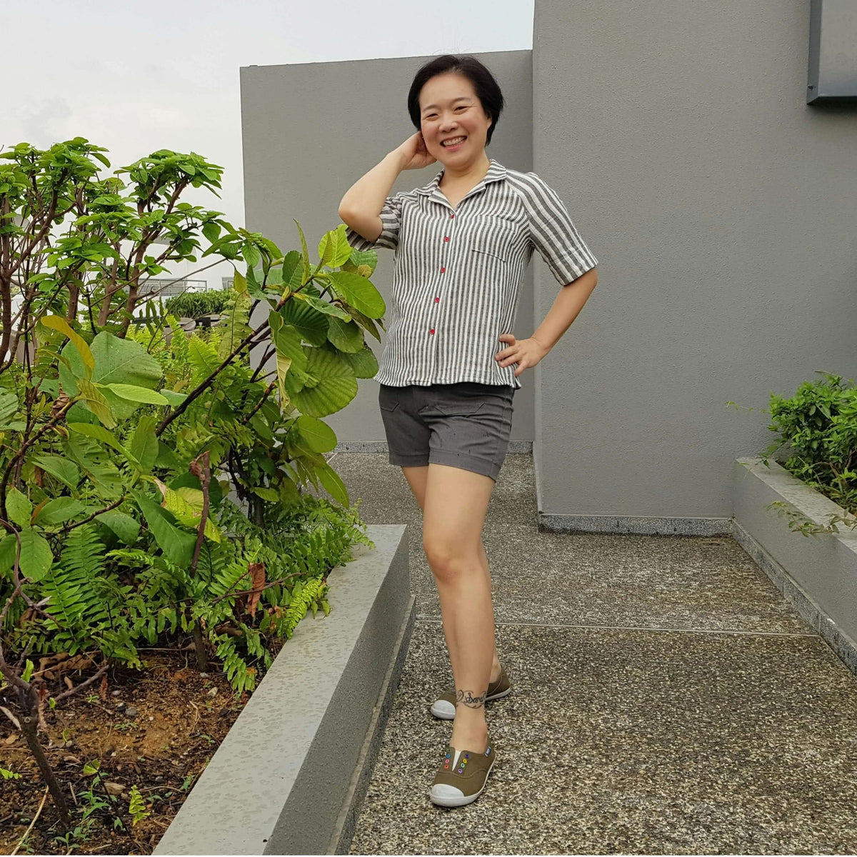 Woman in a Tropicana shirt Paper Pattern outdoors with decorative plants, wearing shorts and sandals, smiling.