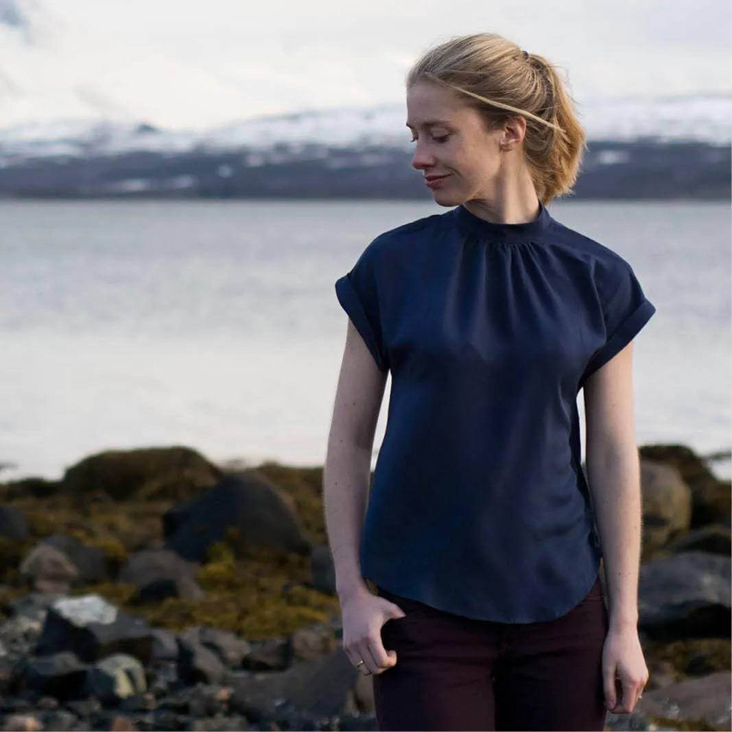 Model wearing Tulip Top Paper Pattern, a cute woven top with gathered neckline and dolman shoulders by a scenic shoreline.