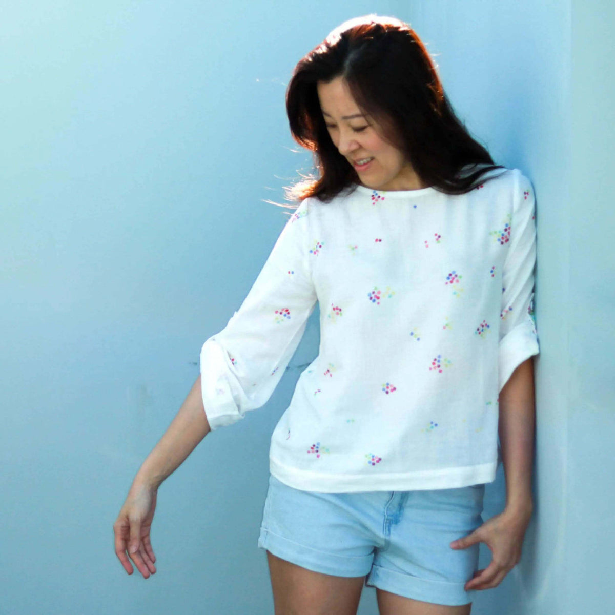 Woman wearing a floral Whisper blouse Paper Pattern with three-quarter sleeves and denim shorts, smiling against a blue background.