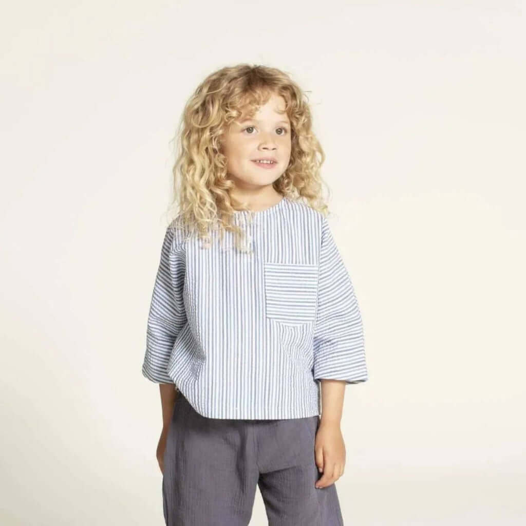 Una shirt sewing pattern model wearing a striped top with front pocket and dolman sleeves for kids size 3-12 years.